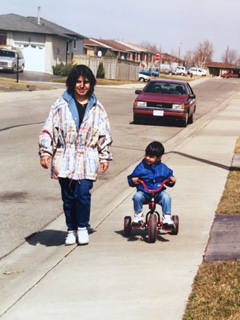 Qasim riding a tricycle along a sidewalk while an adult walks beside him.