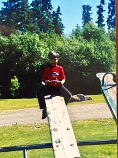 Qasim in a red shirt sitting on a plank in a green park.