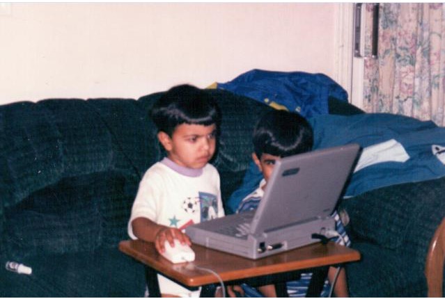 Qasim sitting on a sofa beside another child, both looking at a laptop on a small table.